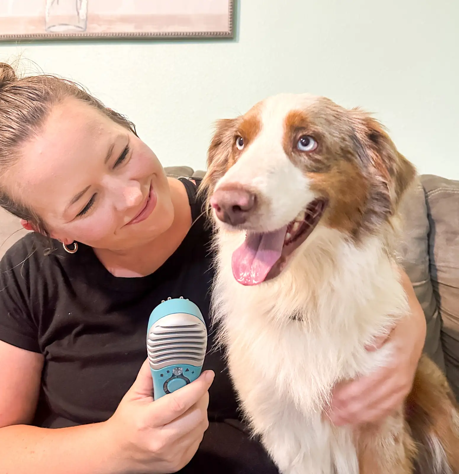 Woman in a black shirt smiles at a fluffy white-and-brown dog while holding a blue microphone on a couch side-by-side pose.