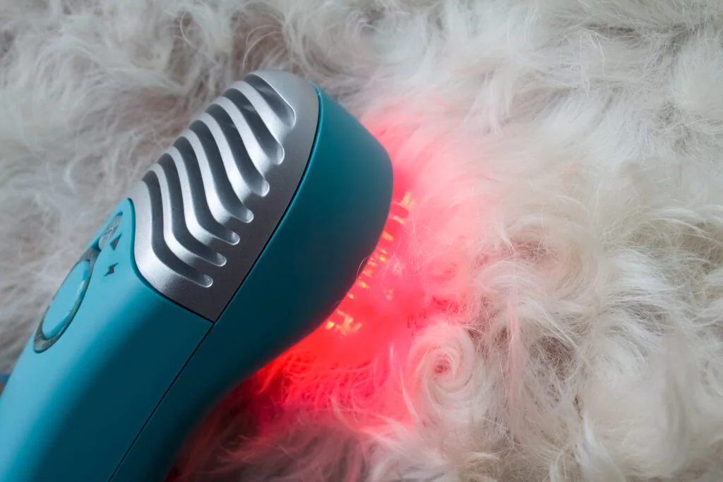 Teal handheld grooming device with a metal comb-like head and red light on fluffy white fur.