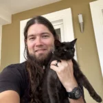 Man with long hair and beard smiles while holding a black cat outdoors on a porch wall behind them.