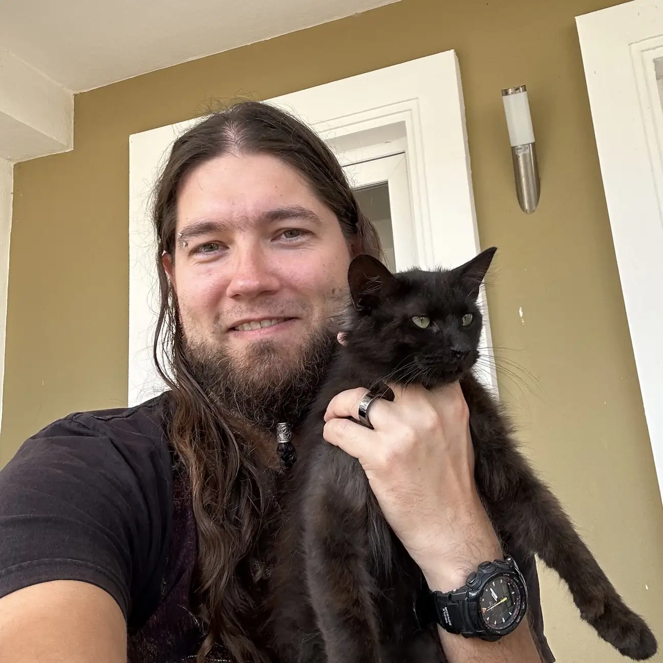 Man with long hair and beard smiles while holding a black cat outdoors on a porch wall behind them.
